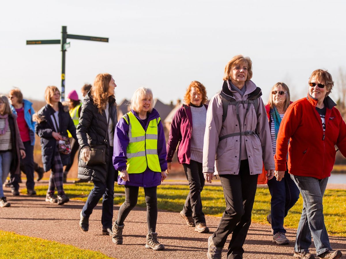 A group of women walking in a parkland scene with a sign post and a lake in the background.