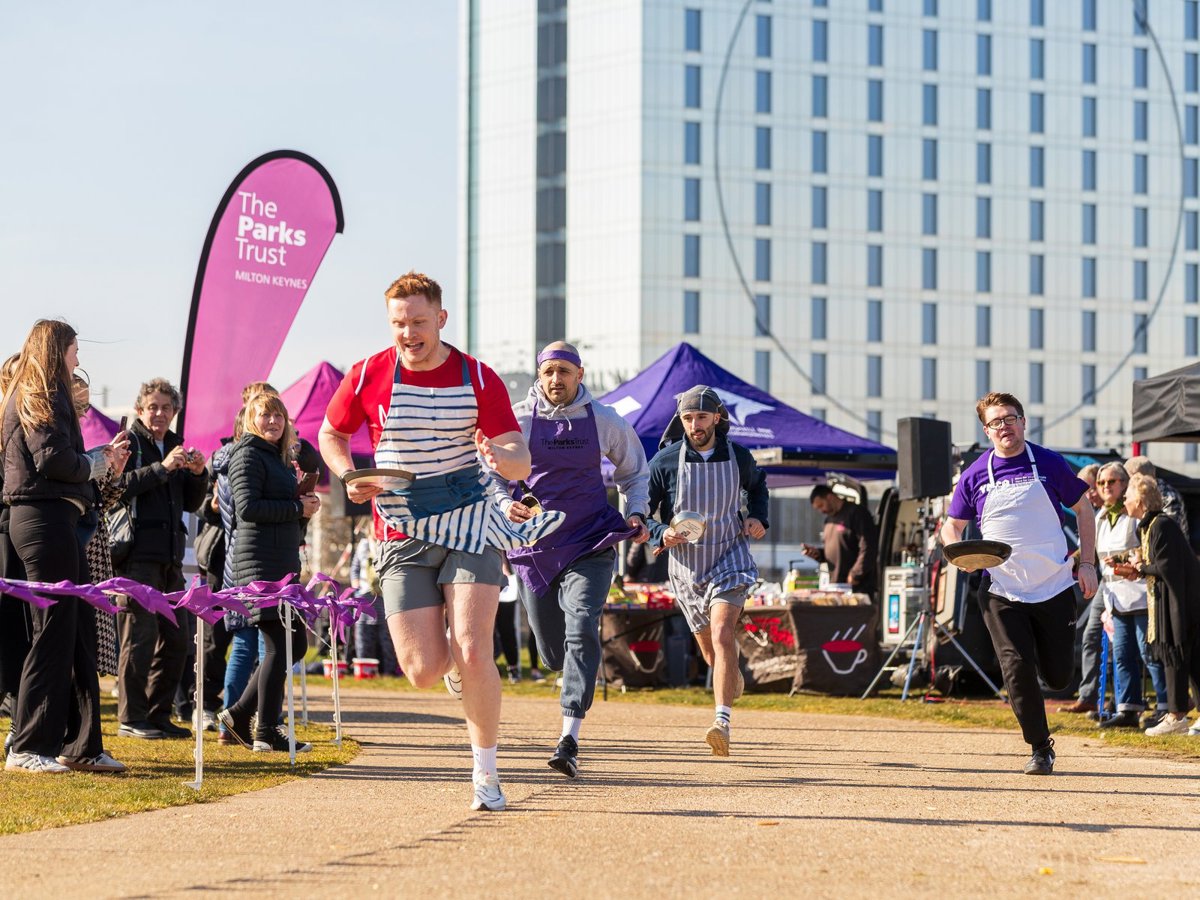 Runners competing in a race holding a frying pan and wearing an apron, with spectators cheering them on