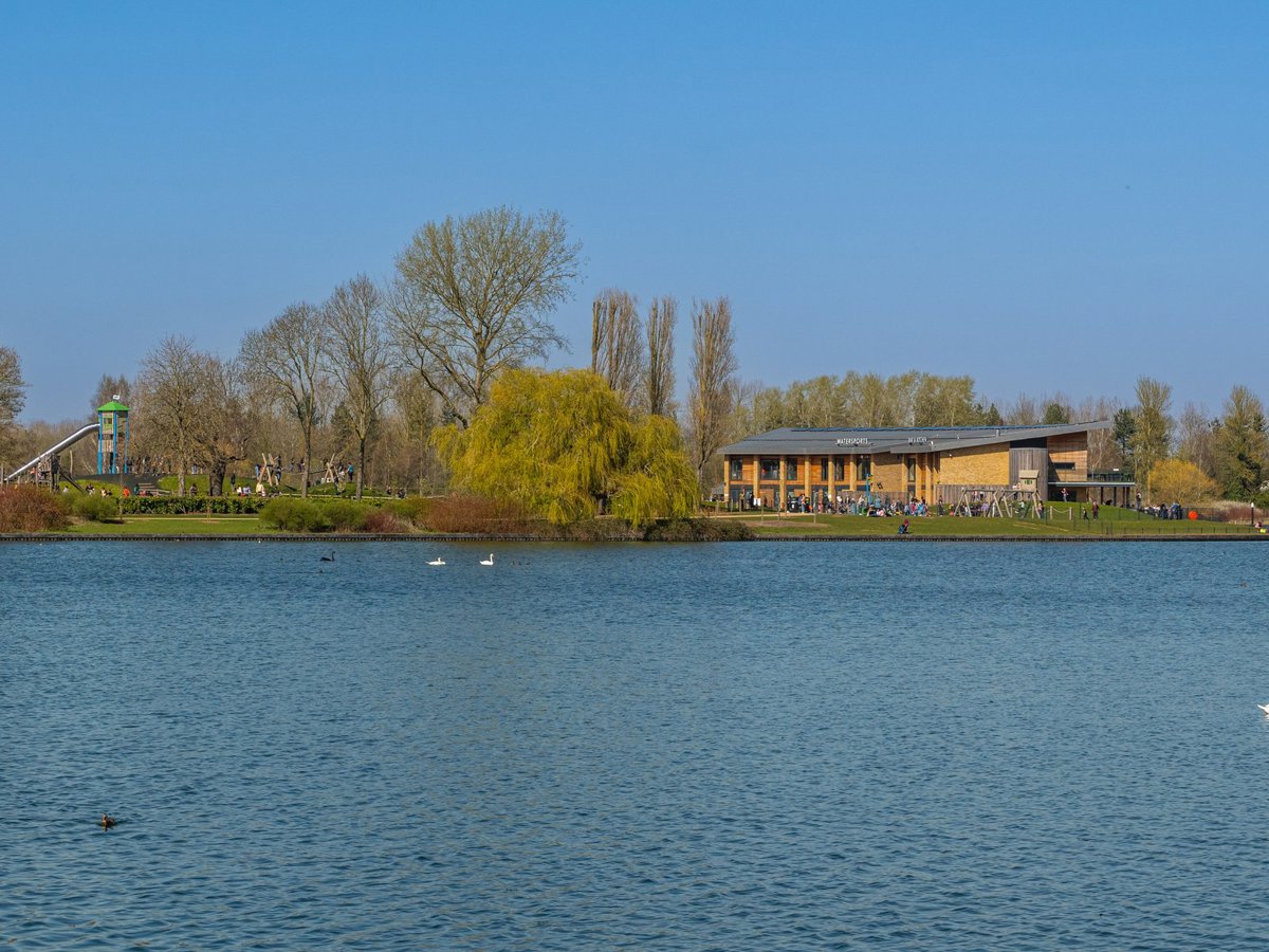 View across Willen Lake looking towards the large water sports centre and outdoor play area on the other side, on a sunny day.